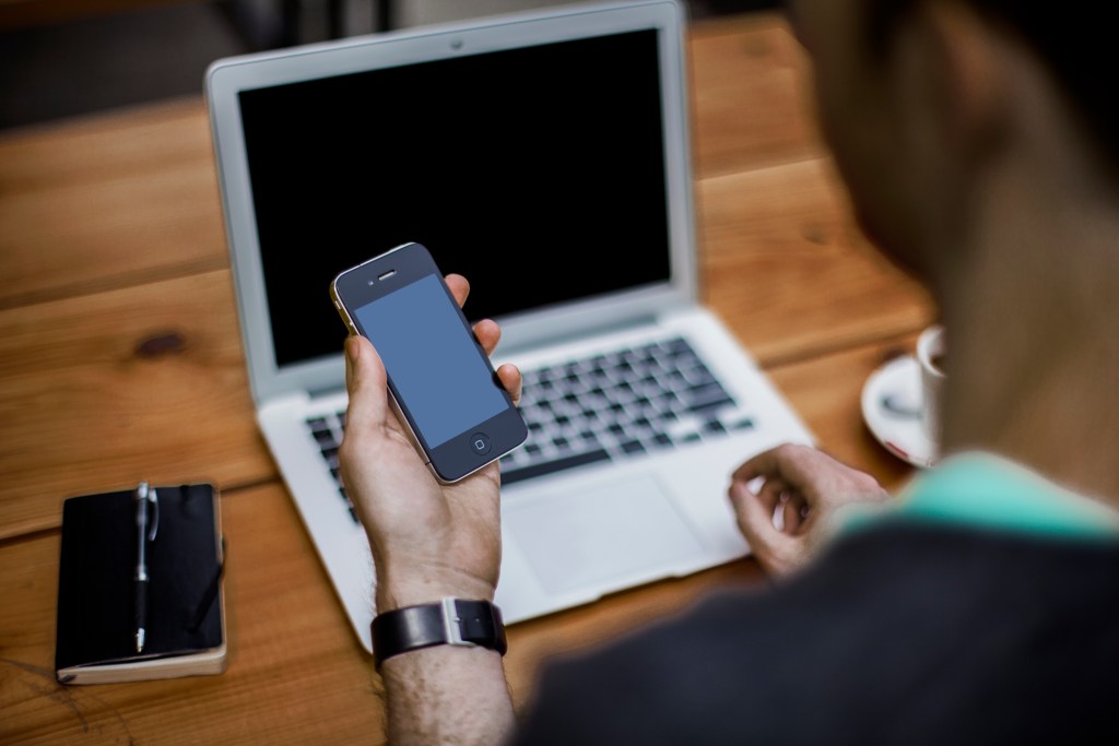 Man using a smartphone beside a laptop at a desk